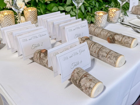 Rustic birch log table place cards on a white tablecloth with lit candle holders and greenery in the background.